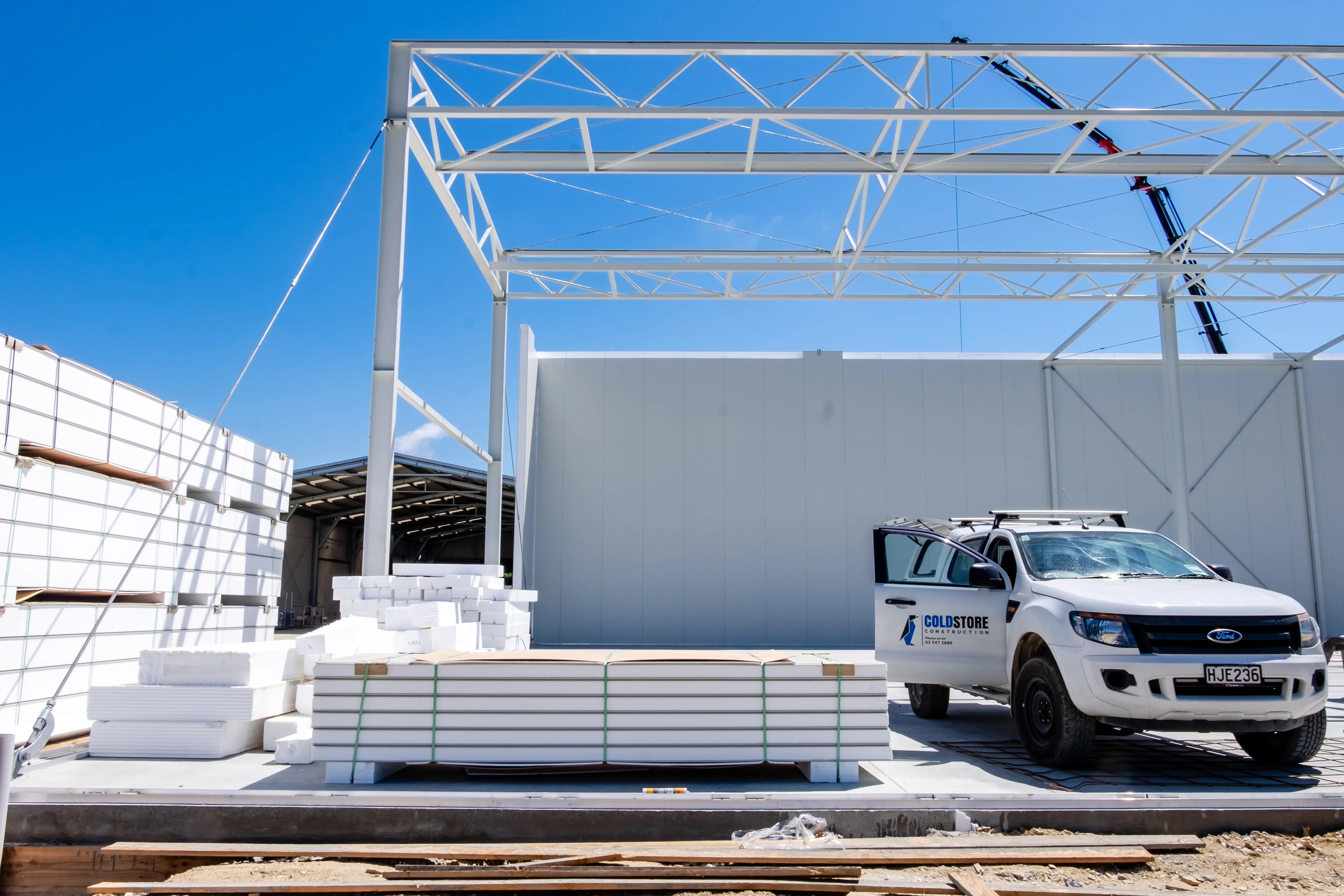 A Coldstore Construction ute on site beside stacked insulated panels and a steel building frame under a clear blue sky.