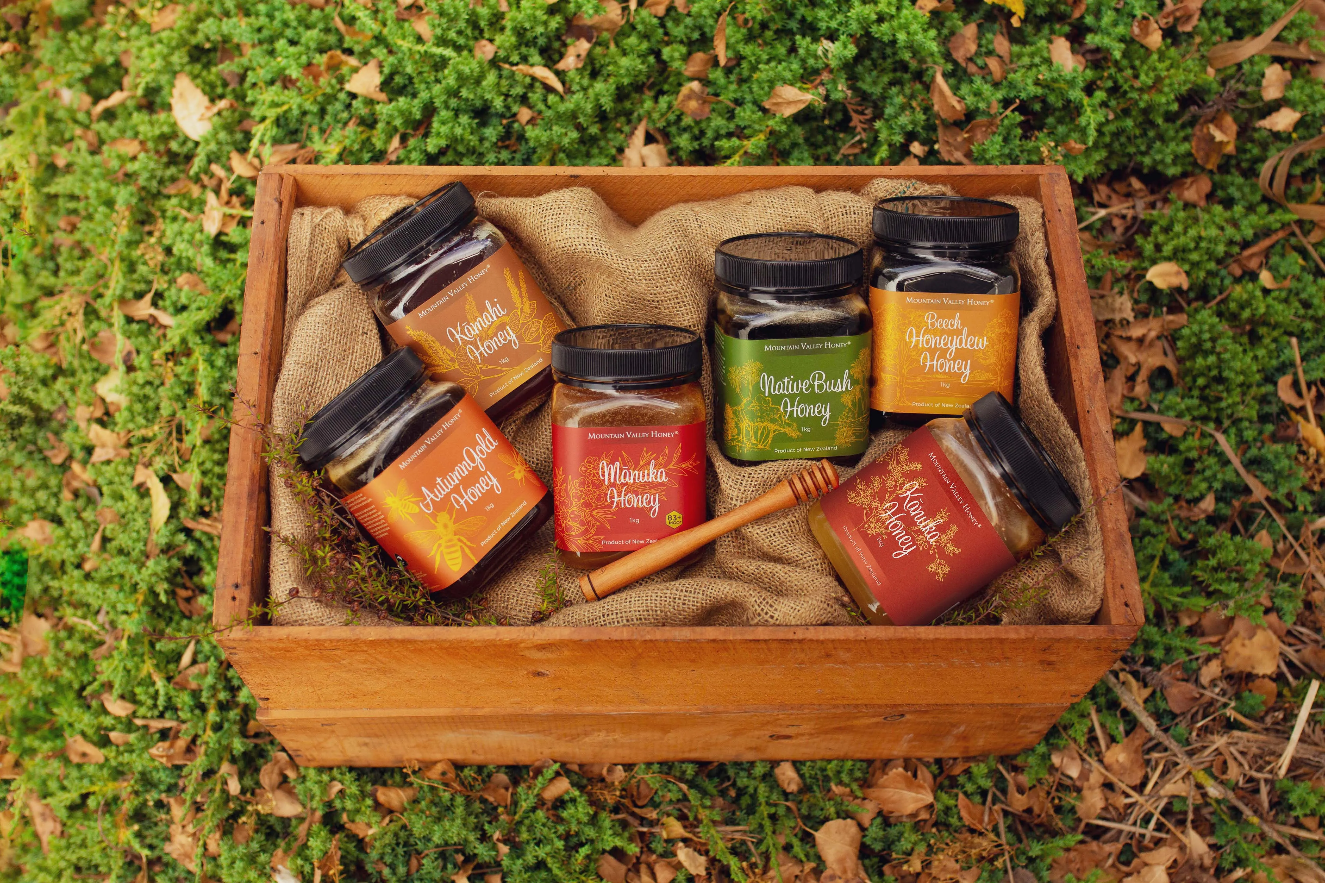 A wooden gift crate of Mountain Valley Honey jars — Mānuka, Native Bush, Kāmahi, Beech Honeydew, Autumn Gold and Kānuka — resting on autumn leaves.